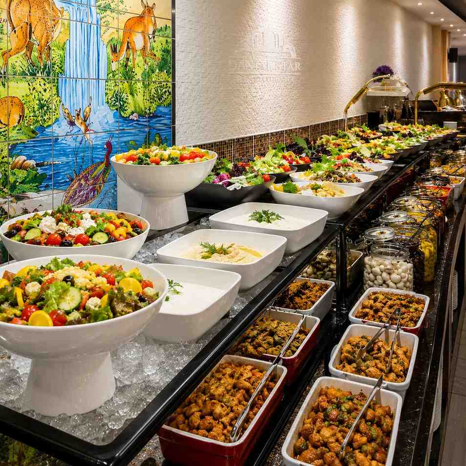 Elegant international buffet display featuring fresh salads, appetizers, and hot dishes arranged along a modern restaurant buffet counter with decorative wall art and warm ambient lighting.